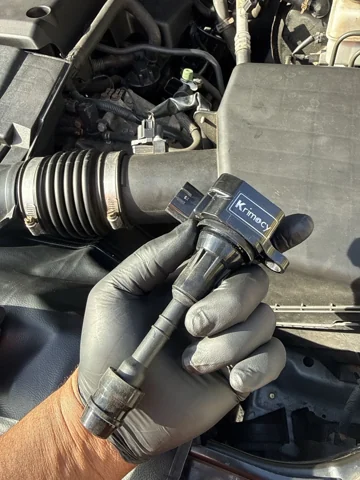 A gloved hand holding an ignition coil beside the engine bay.
